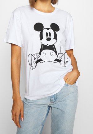 Vrouw draagt een wit t-shirt met een zwart-witte cartoonmuis die met gespreide benen zit, gecombineerd met lichtblauwe spijkerbroek.