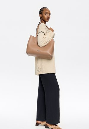 Brown leather tote bag with a smooth texture, wide opening, and subtle hardware accents, carried with a relaxed, oversized cream sweater and dark pants.