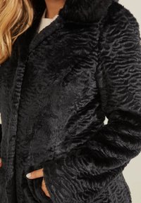 Abrigo de piel sintética negra con patrón texturizado, que presenta un material suave y esponjoso, cuello y bolsillos laterales. Mangas largas con puños anchos.