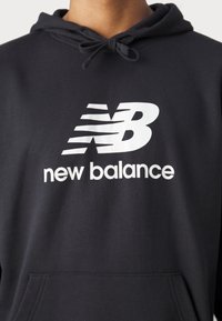 Fekete kapucnis felső, amelyen egy kiemelkedő fehér "New Balance" logó található csíkozott dizájnnal, puha anyagból készült, és elöl zseb található.