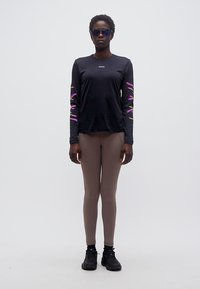 Άτομο που φοράει μαύρο πουκάμισο με μακριά μανίκια και πολύχρωμα σχέδια στα μανίκια, κολάν σε τόνο ταουπέ, μαύρα αθλητικά παπούτσια και ανακλαστικά γυαλιά ηλίου, στέκεται μπροστά σε απλό φόντο.