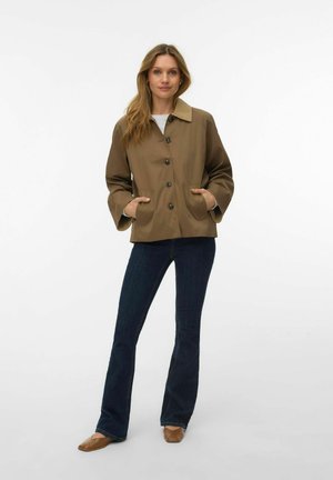 Femme debout portant une veste marron boutonnée, un jean évasé bleu foncé et des chaussures plates marron, sur un fond blanc uni.