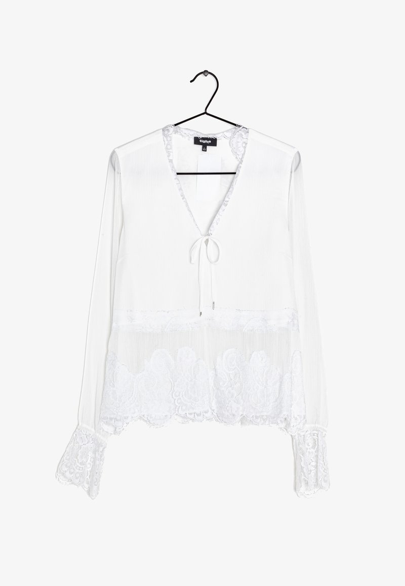 Blouse blanche à manches longues avec finition en dentelle aux poignets et à l’ourlet, col en V avec lien, suspendue sur un cintre noir sur fond blanc.