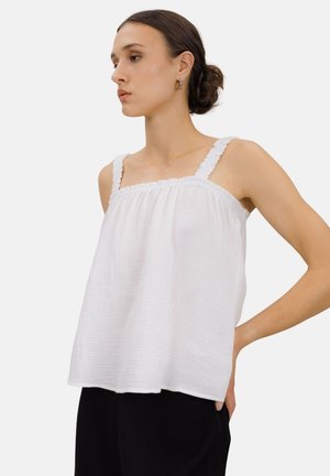 Kvinne med mørkt hår i en lav knute, iført en ermeløs hvit teksturert topp med volangstropper og svarte bukser, stående med hånden på hoften.