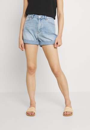 Femme portant un short en denim bleu clair avec ourlet retroussé, un haut noir sans manches et des sandales beige à enfiler, debout devant un fond blanc.