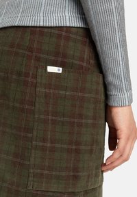 Grön och burgundy rutig kordskjol med sidficka, med en liten etikett. Grå ribbad tröjärm delvis synlig.