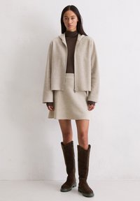 Frau steht vor weißer Wand und trägt eine beigen Reißverschlussjacke, passenden Rock, braunen Rollkragenpullover und dunkelbraune kniehohe Stiefel.