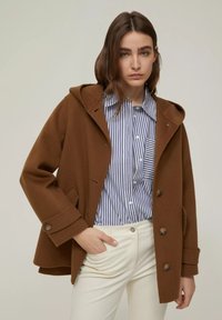 Cappotto in misto lana marrone con cappuccio, bottoni frontali e tasche laterali, indossato sopra una camicia a righe blu e bianca, abbinato a pantaloni bianchi.