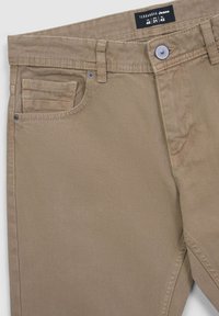Jeans di cotone beige con una superficie liscia, dotati di una tasca frontale, passanti per la cintura e chiusura con bottone metallico. Etichetta del marchio visibile nella parte superiore.