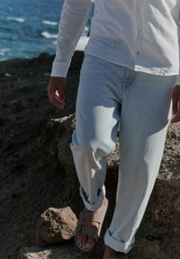 Persona con vaqueros azul claro y sandalias beige caminando por un terreno rocoso cerca del mar, con una camisa blanca de manga larga.