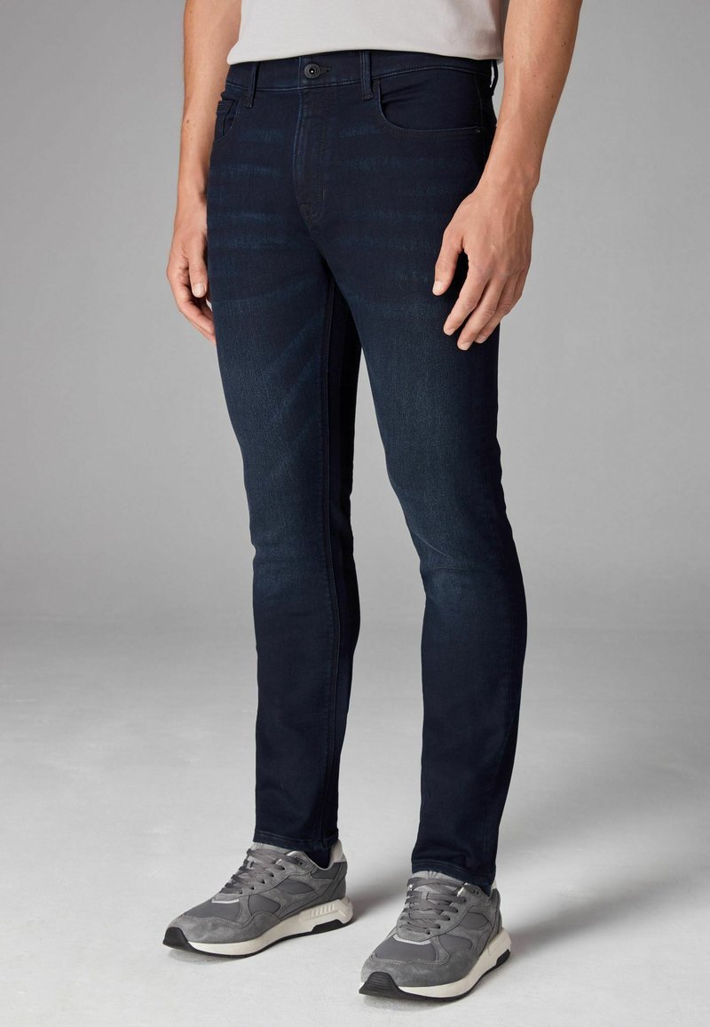 Jeans ajustados de denim azul oscuro, con ligeras variaciones de textura, cinco bolsillos y cierre de botón. Usados con zapatillas grises.