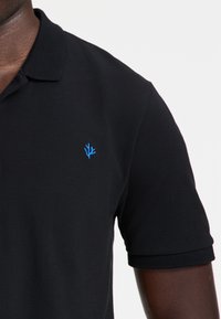 Čierna polokošeľa s textúrovanou tkaninou, krátkymi rukávmi a malým modrým vyšitým logom na ľavej hrudi.