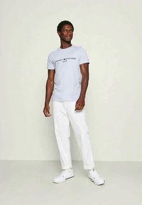 T-shirt de algodão azul claro com o logo "Tommy Hilfiger EST. 1985", combinado com calças brancas e ténis brancos, de pé contra um fundo simples.