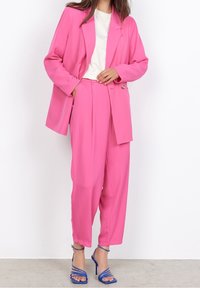 Jasnoróżowy garnitur z dopasowanym żakietem i luźnymi spodniami, zestawiony z białą koszulą i niebieskimi sandałami na obcasie. Gładka, jedwabista faktura.