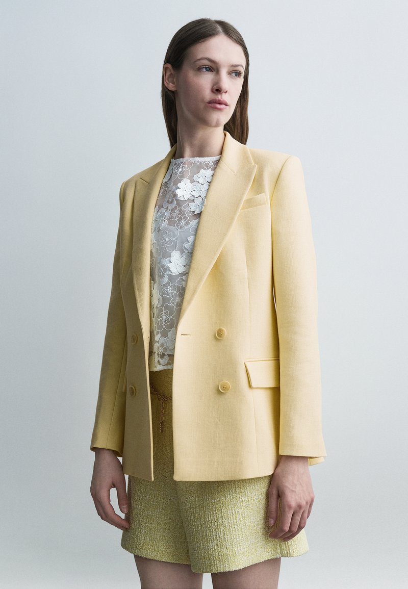 Jeune femme portant un blazer croisé jaune pâle, un haut en dentelle florale blanche et un short jaune texturé, debout devant un fond uni.