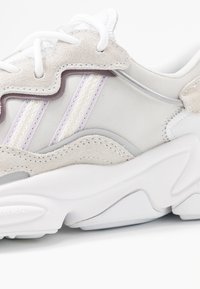 Bílá a béžová semišová teniska s tlustou bílou podrážkou, bílými tkaničkami a dvěma strukturovanými pastelově fialovými pruhy na bočním panelu.