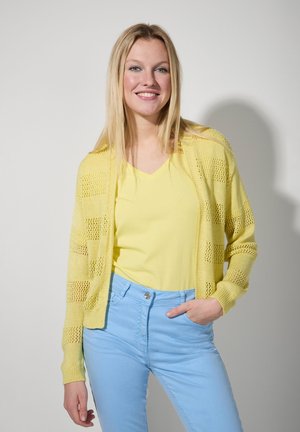 Lichtgele opengehaakte cardigan over een gele V-hals shirt, gecombineerd met lichtblauwe broek. Opvallend gestreept patroon in de cardigan.