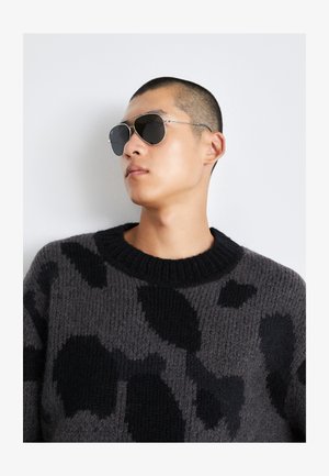 Grå sweater med sort abstrakt mønster, ribbet krave og struktureret strik. Sølvfarvede solbriller med mørke linser, slank rammedesign.