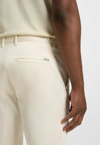 Pantaloni beige chiaro con una texture liscia, dotati di una tasca posteriore e di un accento metallico vicino alla vita. Ingrandimento della mano sul tessuto.