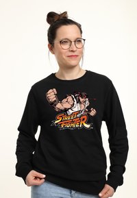 Felpa nera con un'illustrazione grafica di un combattente muscoloso di "Street Fighter" in colori vivaci. Collo rotondo e maniche lunghe.