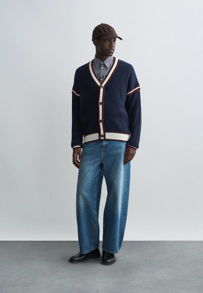 Cardigã tricotado em azul-marinho com riscas brancas e bordô, frente com botões, sobre uma camisa riscada, combinado com jeans de perna larga em azul claro e sapatos pretos.