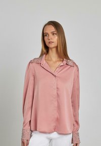 Rosa Satinhemd mit spitzem Kragen, geschmückt mit glänzenden Nieten, langen Ärmeln mit genieteten Manschetten und einem lässigen Schnitt.