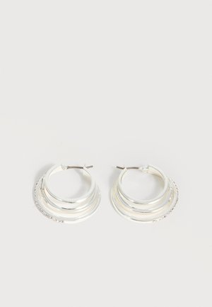 Boucles d'oreilles créoles en argent avec trois bandes semi-circulaires superposées, dont une ornée de petits cristaux clairs sur un fond clair.