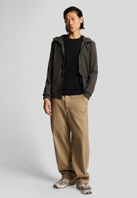 Felpa con zip verde scuro, maglietta nera a girocollo, pantaloni beige a vestibilità morbida e sneakers grigie con dettagli neri. Design semplice e texture liscia.