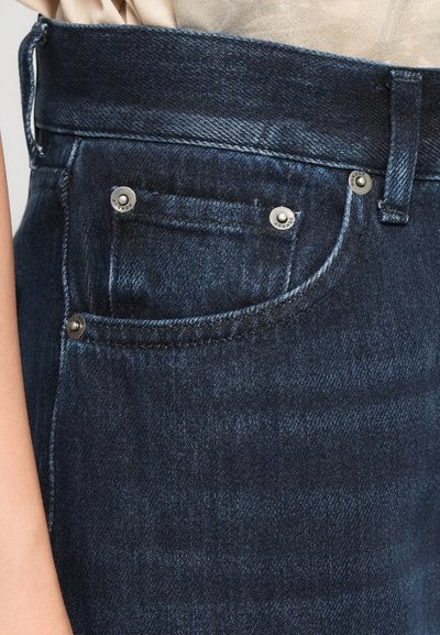 Detailní záběr na tmavě modré denimové džíny s viditelnou přední kapsou, kovovými nýty a poutky na opasek, nošené s béžovým vrškem.
