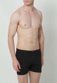 Zwarte katoenen boxershorts met een knoopsluiting, voorzien van een zachte textuur en een elastische tailleband. Model staat met een neutrale achtergrond.