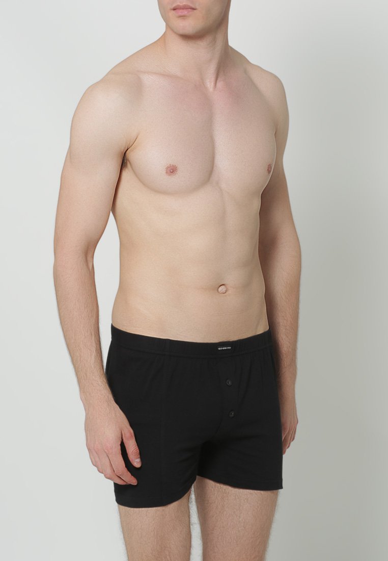 Zwarte katoenen boxershorts met een knoopsluiting, voorzien van een zachte textuur en een elastische tailleband. Model staat met een neutrale achtergrond.