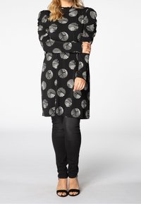 Robe noire avec motif de feuilles blanches, manches longues avec épaules froncées, associée à un pantalon skinny noir et des sandales à talons noires.
