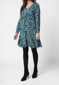 Kukallinen kietaisumekkoティール-värissä mustilla kuvioilla, pitkähihaisilla, V-pääntiellä ja levenevällä helmalla, yhdistetty mustien nilkkureiden kanssa.