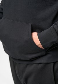 Black hoodie featuring a front pocket, ribbed cuffs, and smooth fabric texture. The model's hand rests inside the pocket.