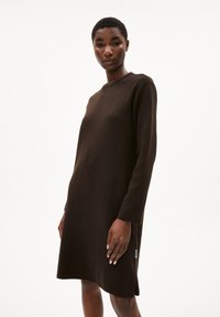 Braunes gestricktes Kleid mit langen Ärmeln, rundem Halsausschnitt und seitlichen Schlitzen. Glatte Textur und lässige Passform. Etikett Detail an der Seite.