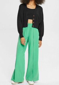 Cardigan nero corto con bottoni in legno abbinato a pantaloni verdi a vita alta e gamba larga, caratterizzati da una texture liscia e dettagli con cuciture a spacco.