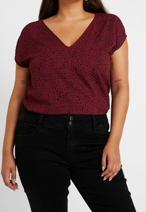 Vrouw draagt een bordeauxrood V-hals top met korte mouwen met een zwart gevlekt patroon en zwarte hooggetailleerde broek, zichtbaar van schouders tot heupen.