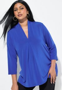 Blusa blu royal con scollo a V, maniche a tre quarti e pieghe frontali, realizzata in un tessuto liscio e morbido. Indossata con pantaloni neri.