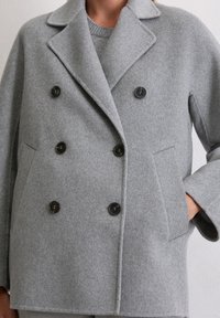 Cappotto di lana grigio doppiopetto con bottoni neri sopra un maglione grigio abbinato, indossato da una persona con una mano nella tasca del cappotto.