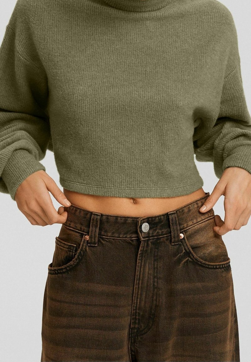 Oříznutý olivově zelený pletený svetr s vysokým límcem, zkombinovaný s vysokým pasem tmavě hnědých džínů z denimu s texturovaným vzorem a zapínáním na knoflíky.