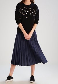 Czarna bluza z metalicznymi detalami, połączona z plisowaną granatową spódnicą midi. Modelka nosi czarne sneakersy.