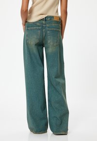 Jeans de pierna ancha en denim verde azulado, con una cintura alta, dos bolsillos traseros y una textura desgastada. La cinturilla tiene una etiqueta de cuero marrón.