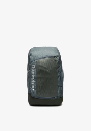Šedý batoh Reebok s velkou přední síťovanou kapsou, boční kapsou na zip a černým dnem, stojící vzpřímeně na bílém pozadí.