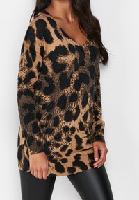 Femme portant un pull en V à manches longues imprimé léopard marron et noir avec un pantalon en cuir noir sur un fond blanc.