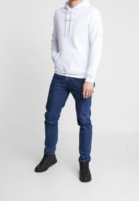 Felpa bianca con cappuccio e tasca frontale, con testo blu. Abbinata a jeans blu scuro e stivali neri con lacci.