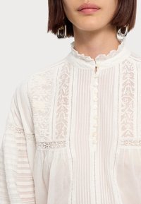 Hvid skjorte med høj flæsekrave, knaplukning foran og detaljeret broderi. Strukturerede striber og blødt stof forbedrer designet.