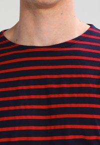 T-shirt blu navy con righe orizzontali rosse, caratterizzato da un collo rotondo e maniche corte. Il tessuto appare morbido con una leggera elasticità.