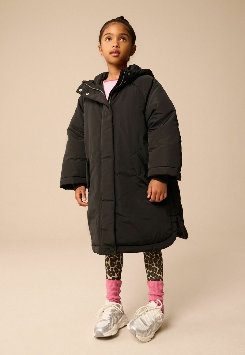 Cappotto imbottito nero con cappuccio, tasche frontali e chiusura a pressione. Indossato sopra un top rosa, leggings a stampa leopardata e sneakers argentate con calzini rosa.