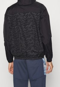 Svart vindjacka med ett vågigt grått mönster på baksidan, elastiska ärmslut och dragsko i nederkant för justerbar passform.