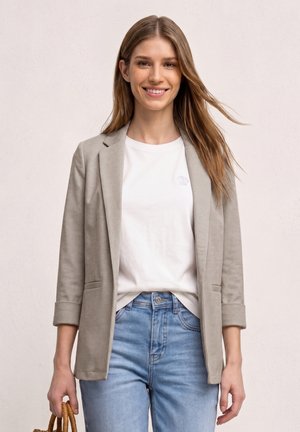 Jeune femme souriante, portant un blazer beige sur un t-shirt blanc et un jean bleu clair, tenant un petit sac à main rond tissé.
